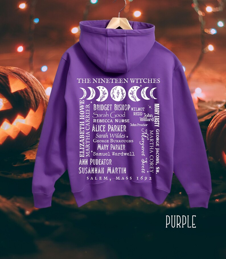 Op de afbeelding: Een paarse hoodie met een witte afbeelding op de achterkant. Het ontwerp bevat maanfasen, de tekst THE NINETEEN WITCHES, SALEM, MASS 1692 en een lijst met namen waaronder Bridget Bishop, Sarah Good, Rebecca Nurse en anderen.