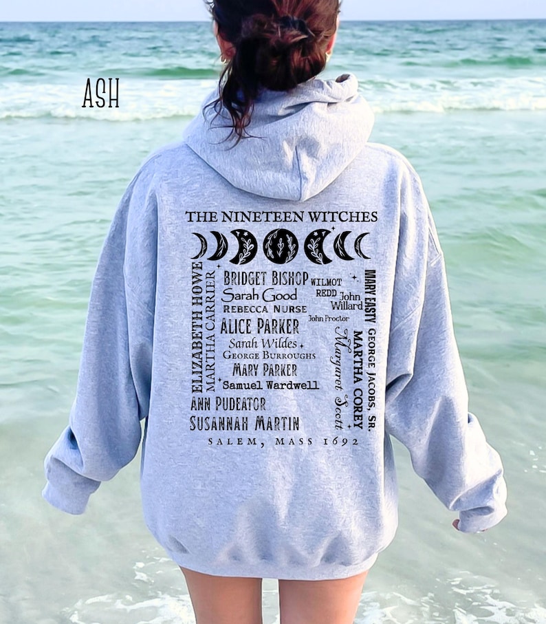 Op de afbeelding: Een asgrijze hoodie met een zwarte opdruk op de achterkant. Het ontwerp bevat maanfasen, de tekst THE NINETEEN WITCHES, een lijst met namen waaronder Bridget Bishop en Sarah Good, en de tekst SALEM, MASS 1692 onderaan het ontwerp.