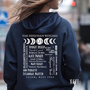 Op de afbeelding: Een marineblauwe hoodie met een witte afbeelding op de achterkant. Het ontwerp bevat de tekst THE NINETEEN WITCHES, een rij maanfasen en een lijst met namen waaronder Bridget Bishop en Sarah Good, eindigend met SALEM, MASS 1692.