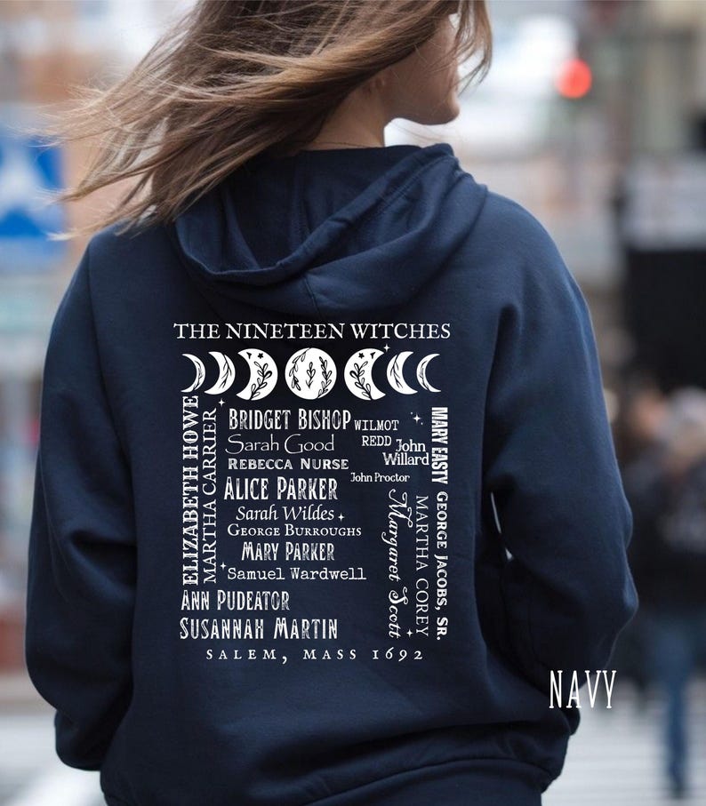 Op de afbeelding: Een marineblauwe hoodie met een witte afbeelding op de achterkant. Het ontwerp bevat de tekst THE NINETEEN WITCHES, een rij maanfasen en een lijst met namen waaronder Bridget Bishop en Sarah Good, eindigend met SALEM, MASS 1692.