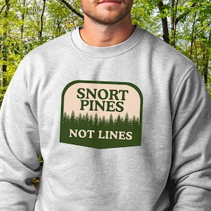 May include: A light gray sweatshirt with a green and tan graphic that reads "SNORT PINES NOT LINES". The graphic features a forest of pine trees. The sweatshirt is a crew neck style.