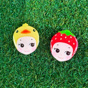 Op de afbeelding: Twee decoratieve gezichtsvormige spelden. Eén is geel met een eendenontwerp, de andere is rood met een aardbeienontwerp. Beide hebben lachende gezichten met zwarte ogen en roze wangen. De spelden zijn geplaatst tegen een groene, grasachtige achtergrond.