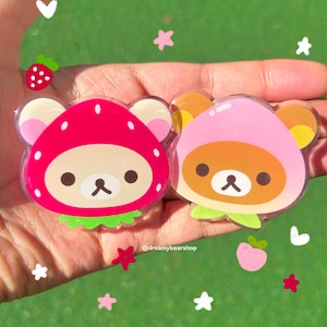 May include: Two clear plastic charms featuring a cartoon bear wearing a strawberry and peach hat. The charms are on a hand against a green background with white stars and hearts.