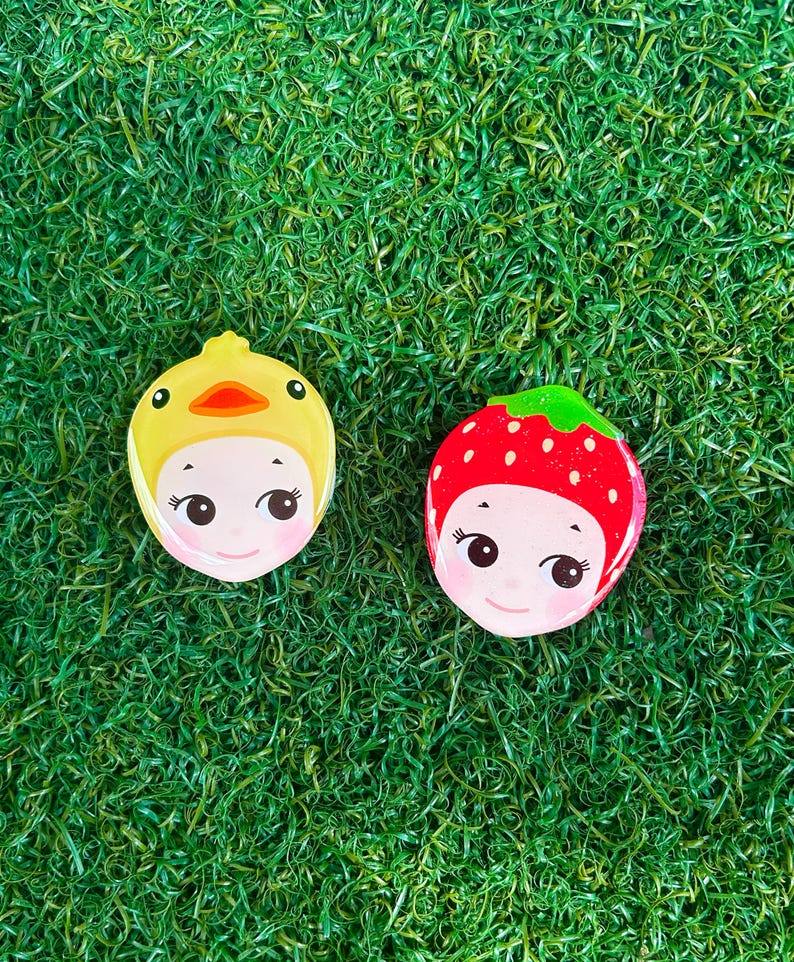 May include: Two decorative face-shaped pins. One is yellow with a duck design, the other is red with a strawberry design. Both have smiling faces with black eyes and pink cheeks. The pins are set against a green, grassy background.