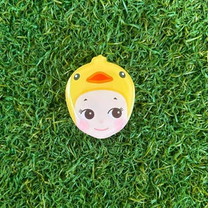 May include: A yellow, duck-shaped compact mirror with a cartoon face. The mirror features a yellow duck hood with an orange beak and black eyes. The face has pink cheeks and is set against a backdrop of green grass.