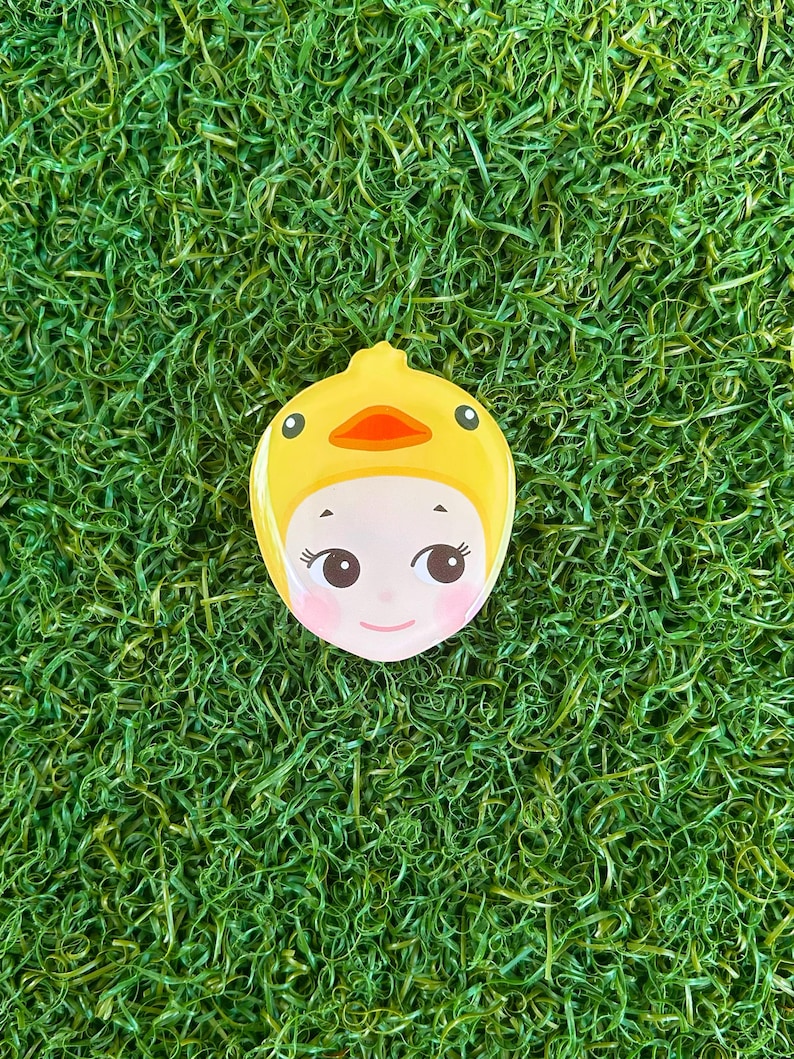 May include: A yellow, duck-shaped compact mirror with a cartoon face. The mirror features a yellow duck hood with an orange beak and black eyes. The face has pink cheeks and is set against a backdrop of green grass.