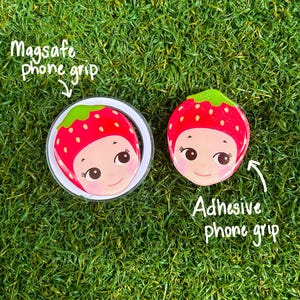 May include: Two phone grips with a strawberry design, one Magsafe and one adhesive. The strawberry design features a smiling face with a red body, green top, and white border. The text "Magsafe phone grip" and "Adhesive phone grip" are also visible.