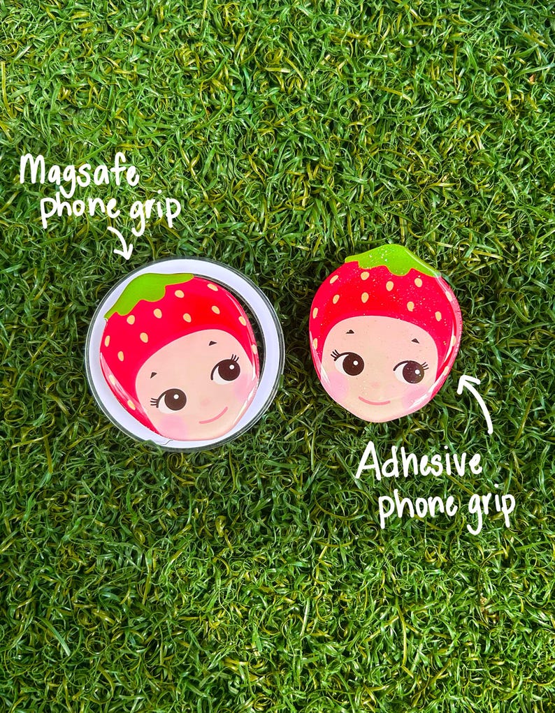 May include: Two phone grips with a strawberry design, one Magsafe and one adhesive. The strawberry design features a smiling face with a red body, green top, and white border. The text "Magsafe phone grip" and "Adhesive phone grip" are also visible.