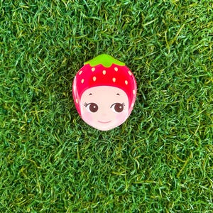 May include: A whimsical, red strawberry-shaped compact mirror with a green stem and a smiling face. The mirror has a glossy finish and is set against a backdrop of vibrant green artificial grass.