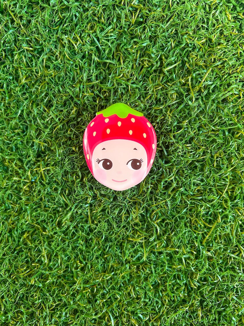 May include: A whimsical, red strawberry-shaped compact mirror with a green stem and a smiling face. The mirror has a glossy finish and is set against a backdrop of vibrant green artificial grass.