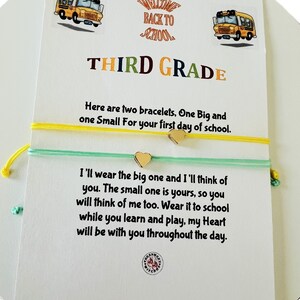Puede incluir: Una tarjeta con fondo amarillo y verde y el texto "WELCOME BACK TO SCHOOL THIRD GRADE" con dos ilustraciones de autobuses escolares. La tarjeta tiene un mensaje sobre dos pulseras, una grande y una pequeña, con un dije de corazón en cada una.