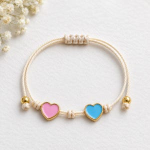 Puede incluir: Una pulsera de cordón color crema con dos dijes en forma de corazón con bordes dorados, uno relleno de esmalte rosa y el otro de esmalte azul claro, acentuada por dos pequeñas cuentas doradas en los extremos del cordón ajustable.