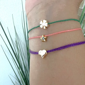 May include: Three friendship bracelets with different charms. The top bracelet is green with a four-leaf clover charm. The middle bracelet is pink with a star charm. The bottom bracelet is purple with a heart charm.