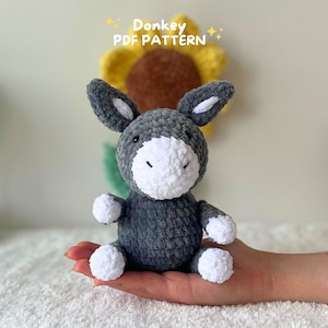 May include: A gray crocheted donkey toy with white crocheted details. The donkey is sitting in a person's hand. The background is a yellow and brown crocheted sunflower.