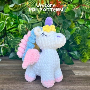 Puede incluir: Un unicornio de crochet blanco con alas rosas, una crin azul y rosa y un cuerno amarillo y morado. El unicornio está de pie sobre una superficie de madera. "Unicorn PDF Pattern" está escrito en el fondo.
