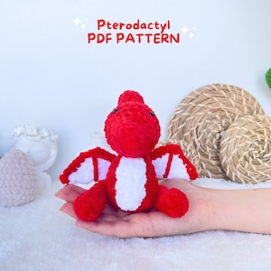 May include: A red and white crocheted Pterodactyl amigurumi toy. The toy is sitting on a hand. The text "Pterodactyl PDF PATTERN" is above the toy.