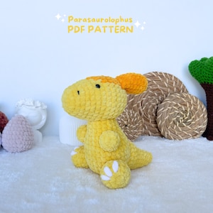 May include: A crocheted yellow Parasaurolophus dinosaur plush toy with a bright orange crest. The toy is sitting on a white fluffy surface with a wicker basket and a small white ceramic pot in the background.