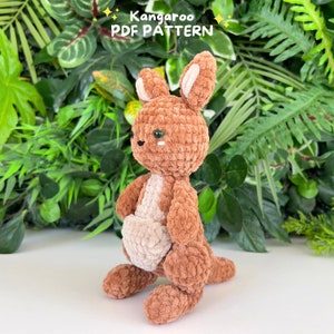 May include: A brown crocheted kangaroo plush toy with a white pouch. The toy has green eyes and is standing on a white surface. The text "Kangaroo PDF PATTERN" is visible above the toy.