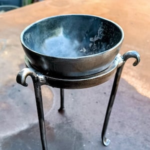 May include: A small, round, metal bowl sits on a three-legged stand. The bowl and stand are both a dark, metallic grey colour.