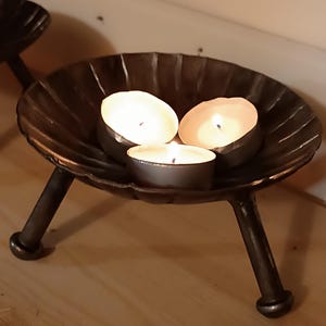Puede incluir: Tres velas votivas están encendidas en un candelabro de metal negro con tres patas. El candelabro está sobre una superficie de madera.