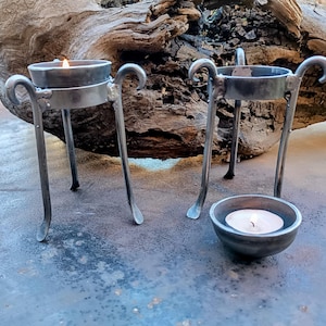 May include: Two black metal candle holders with three legs each. The candle holders are made of wrought iron and have a rustic look. Each holder has a small, round candle dish on top. One candle is lit and the other is not.