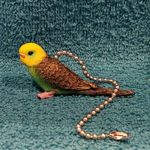 May include: A decorative budgie figurine with a yellow head, green body, and brown wings. The bird has a silver ball chain attached. The figurine is resting on a teal, textured surface.