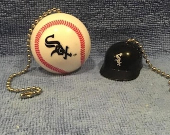 Pode incluir: Um conjunto com tema de beisebol com uma bola de beisebol branca com costuras vermelhas e o logotipo do Chicago White Sox, e um capacete de beisebol preto em miniatura com o logotipo da equipe. Ambos os itens têm uma corrente de bolinhas prateada.