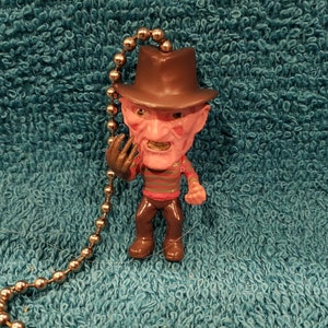 May include: A small, detailed figurine of a character with a brown hat, red skin, and a striped shirt. The figurine is attached to a metal ball chain with a cylindrical pull. The background is a textured teal fabric.