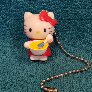 May include: A white Hello Kitty figurine with a red bow, wearing a red dress, holding a bowl of yellow food and a spoon. A silver ball chain is attached. The figurine is on a teal background.