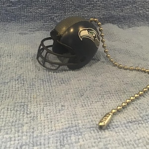 May include: A black and green football helmet with a white logo of a bird's head. The helmet is attached to a gold chain.