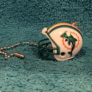 May include: Miniature white football helmet with green and orange accents, featuring the Miami Dolphins logo. The helmet has a green face mask and is attached to a silver ball chain. The helmet is a collectible item.