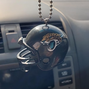 May include: A miniature black football helmet hanging from a silver ball chain. The helmet features a jaguar head logo on the side, depicted with gold, black, and teal colors, showing an open mouth with a teal tongue and sharp white teeth.