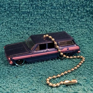 May include: A navy blue toy station wagon with pink and blue accents, and a silver ball chain. The miniature car has black wheels with blue rims. The car is resting on a teal, textured surface.