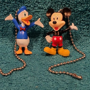 May include: Two Disney character fan pull chains. Mickey Mouse is wearing his signature red shorts and yellow shoes. Donald Duck is wearing a blue sailor shirt with a red bow tie and yellow shoes.