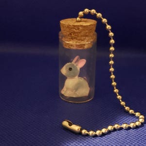 May include: A small glass bottle with a cork stopper, containing a miniature rabbit figurine. The rabbit is light blue with pink ears. A silver ball chain is attached to the cork. The background is a solid dark blue.