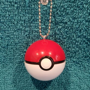 May include: A red and white Poké Ball with a black stripe and a white button. The ball is attached to a silver ball chain and a clear suction cup. The background is a textured teal fabric.