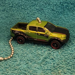 May include: A green and black Ford Ranger toy truck with red-rimmed wheels. The toy truck has a black hood and a silver ball chain attached. The word "Ford" is visible on the truck.
