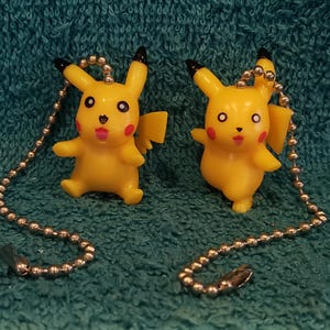 May include: Two yellow Pikachu character figures, each attached to a silver-toned metal ball chain. The figures feature pointed ears with black tips, red circular cheeks, and a lightning bolt-shaped tail. Both figures have a smooth plastic finish.