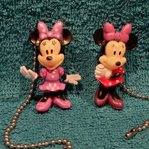 May include: Two plastic Minnie Mouse figurines with pull chains. The figurines are wearing pink and red dresses with white polka dots and have pink bows in their hair.