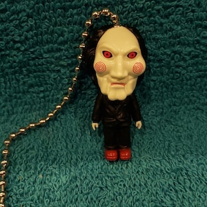 May include: A white plastic figurine of a horror movie character with a red spiral pattern on the face. The figurine is attached to a silver chain.