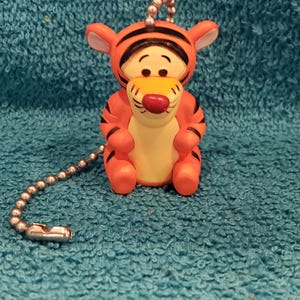May include: Orange and black Tigger figurine with a silver chain. The figurine has a yellow belly, a red nose, and black stripes. The Tigger figurine is a character from the Winnie the Pooh series.