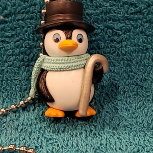 May include: A charming penguin figurine wearing a black top hat, a light blue scarf, and holding a silver cane. The penguin has a white belly, black back, orange feet and beak, and blue eyes. It is attached to a silver ball chain.
