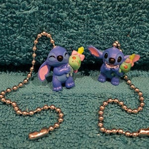 May include: Two blue Stitch figurines, each holding a green and yellow treat with a pink bow. The figurines are attached to silver ball chains. The background is a textured teal surface.