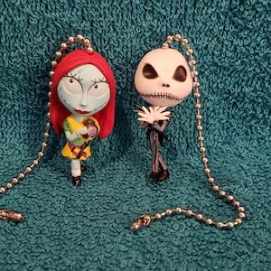 May include: A pair of Nightmare Before Christmas fan pull chains featuring Jack Skellington and Sally. Jack is wearing his signature black and white suit and Sally is holding a jar of eyeballs. The chains are silver with small round beads.