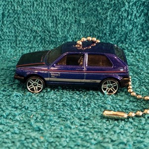 May include: A die-cast model car in dark blue with a black roof. The car has silver wheels and red and white stripes. A silver chain is attached to the car.