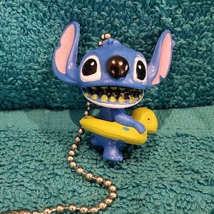 May include: A blue Stitch figurine with large ears, black eyes, and a wide grin, holding a yellow inflatable ring. The figurine is attached to a silver ball chain, likely for use as a pull chain or keychain.