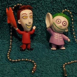 May include: Two Nightmare Before Christmas light switch pull chains. One features a red-clothed character with a red and orange wing. The other features a purple-clothed character with a white cup.