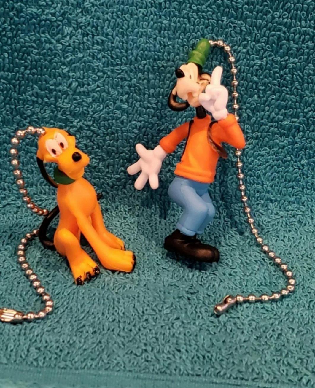 May include: A set of two plastic fan pull chains featuring cartoon characters. The first chain features Goofy wearing a green hat and an orange shirt. The second chain features Pluto, a yellow dog, sitting down. Both chains are silver.