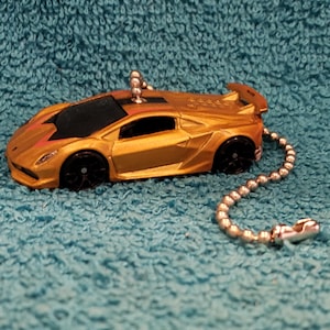 May include: A gold-colored toy car with black wheels and a silver chain. The car has a spoiler and a sleek design. The keychain is on a blue textured background.