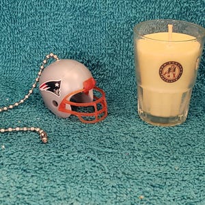 May include: A small, white football helmet with a red face mask and a New England Patriots logo. The helmet is attached to a silver chain. A clear glass jar with a yellow candle inside. The candle has a New England Patriots logo on the side.
